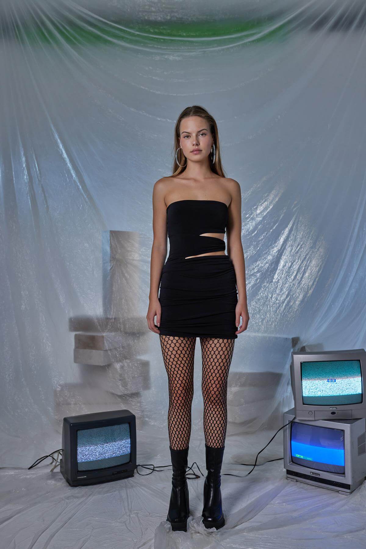 Model wearing a black pleated mini bodycon dress with fishnet stockings, posed in a stylized indoor setting.
