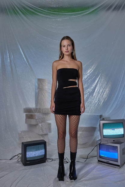 Model wearing a black pleated mini bodycon dress with fishnet stockings, posed in a stylized indoor setting.